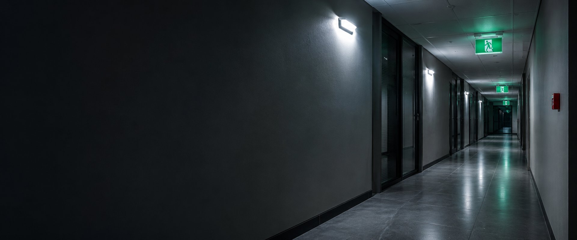 Darkened commercial corridor at night lit by an emergency LED bulkhead with a green SANS running-man exit sign at the far end